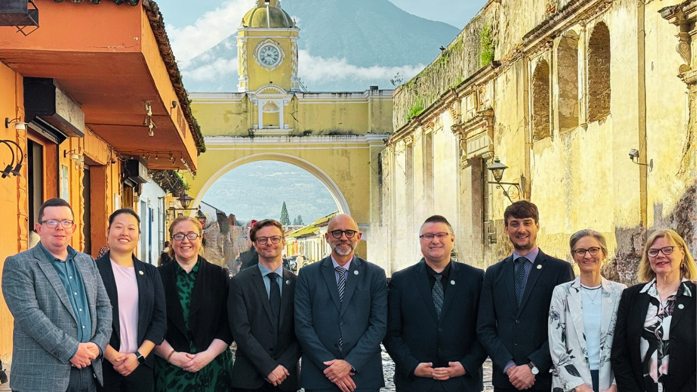 A group of nine people in business attire stand in front of the Santa Catalina Arch in Antigua, Guatemala, with a volcano visible in the background.