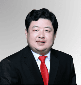 A man wearing a black suit, white shirt, and red tie poses in front of a plain gradient background.