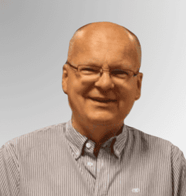 A middle-aged man with glasses, a bald head, and a striped button-up shirt smiles at the camera against a light grey background.