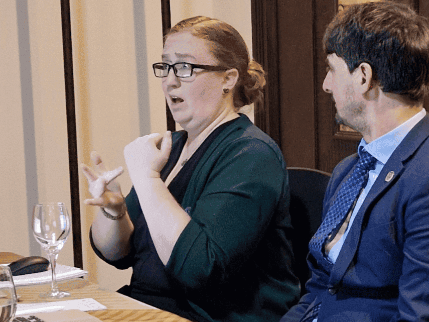 A woman in glasses uses sign language while seated at a conference table next to a man in a suit. Laptops, documents, and water glasses are on the table.