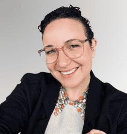Person with short, curly hair and glasses, wearing a dark blazer and beaded necklace, smiling at the camera against a light grey background.