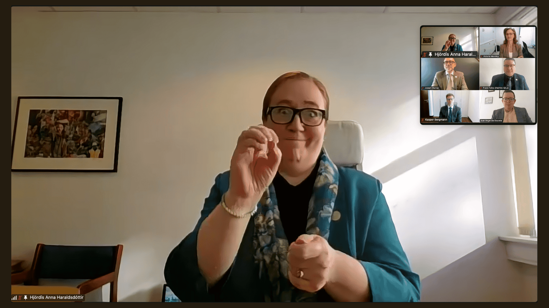 A woman uses sign language during a video conference; five other participants are visible in small windows in the top-right corner of the screen.