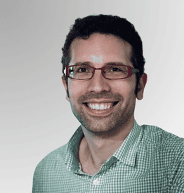 A man with short dark hair, glasses with red frames, and a green chequered shirt smiles against a light gradient background.