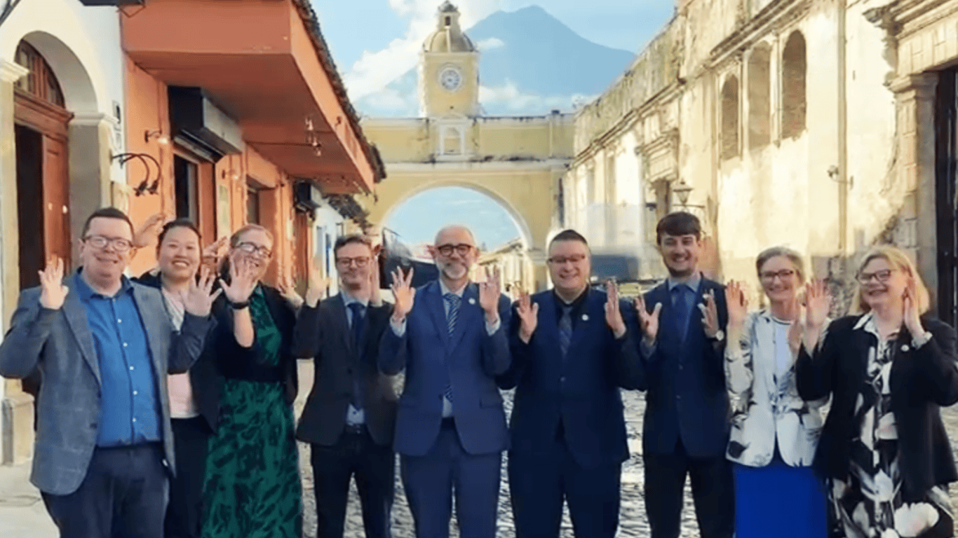 A group of nine people in formal attire stand on a cobblestone street, smiling and holding up their hands, with an arched clock tower and a mountain in the background.