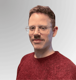 A man with short, light brown hair, glasses, and a moustache is wearing a maroon jumper and facing the camera against a grey gradient background.