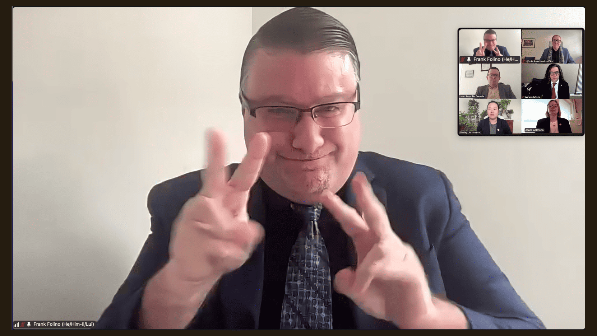 A man in a suit and glasses signs with both hands during a video call; four other participants appear in smaller windows in the top right corner.