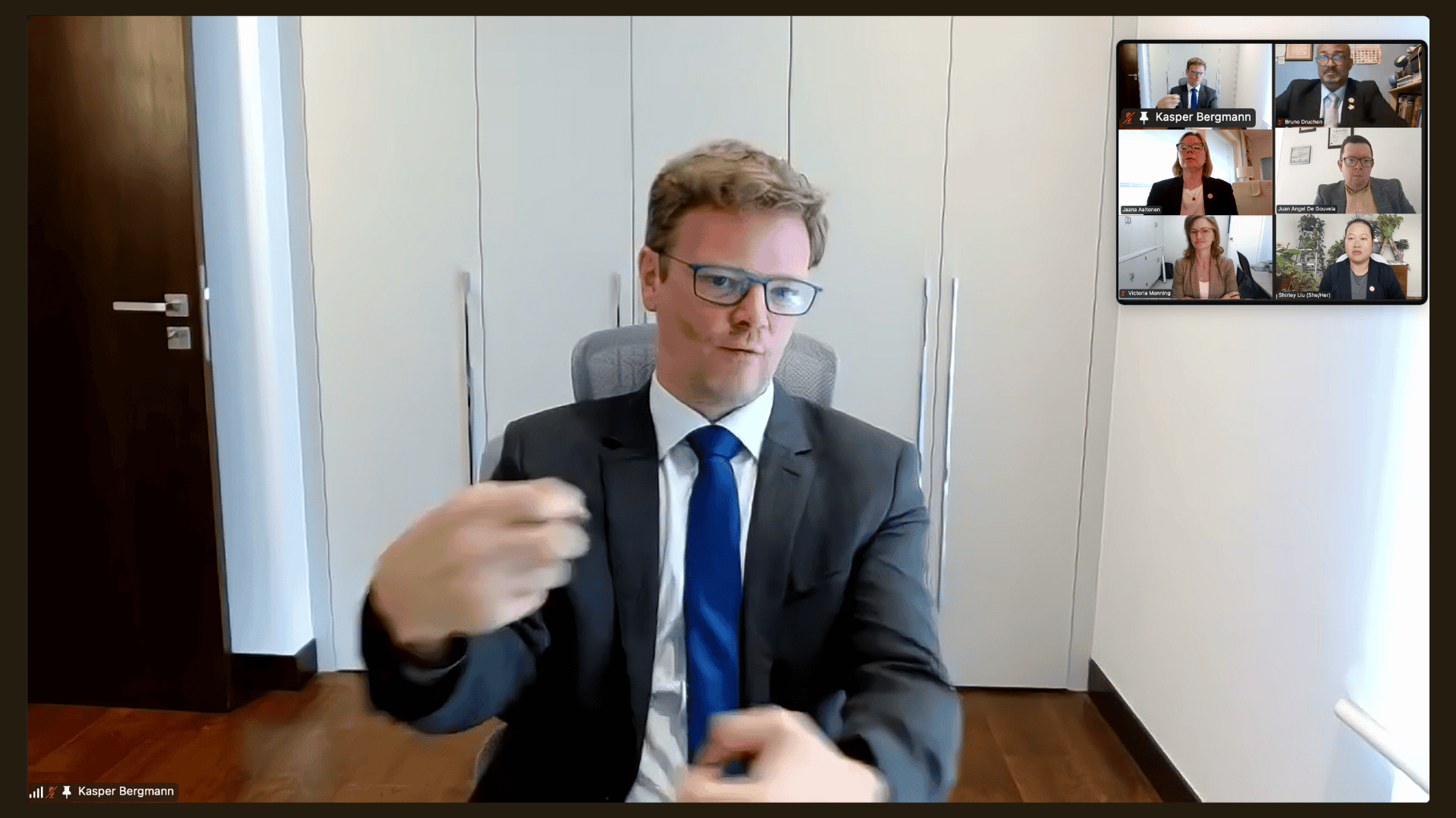 A man in a suit sits in a modern office, gesturing with his hands during a video conference with five participants visible in a sidebar.