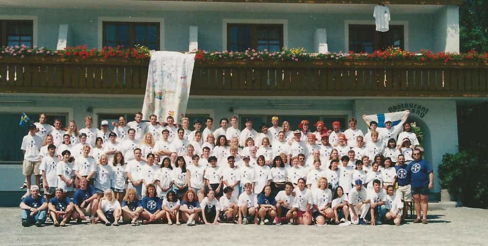 A large group of people wearing matching white shirts pose together in front of a building with a wooden balcony decorated with flowers.