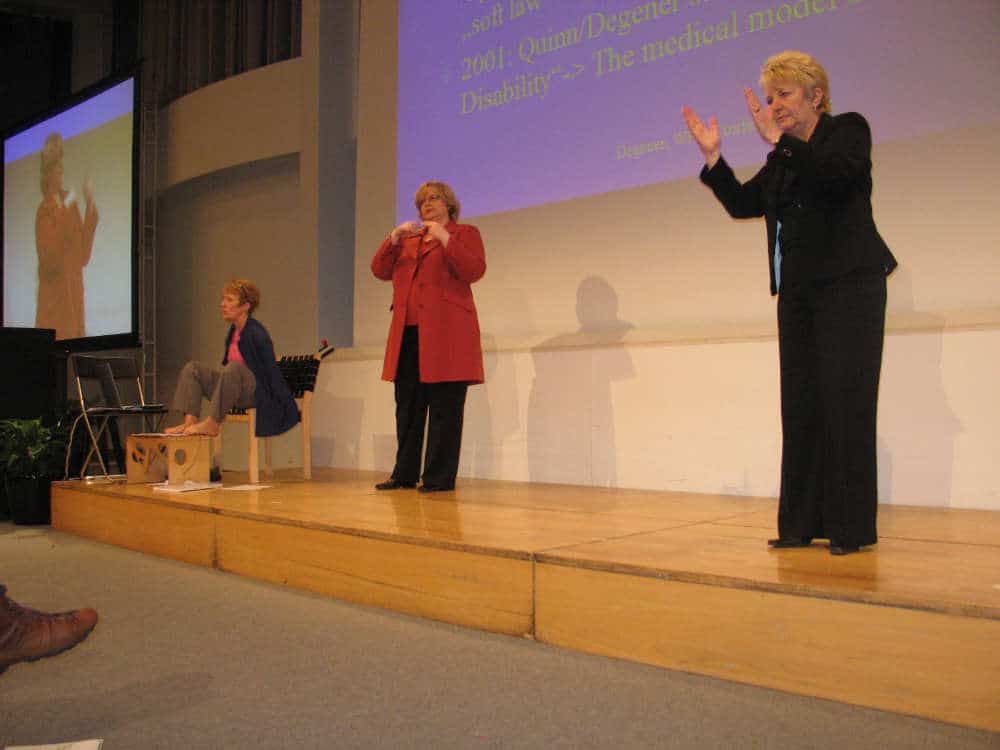 Three women are on stage: one is seated, one is signing, and one is speaking near a screen displaying text.
