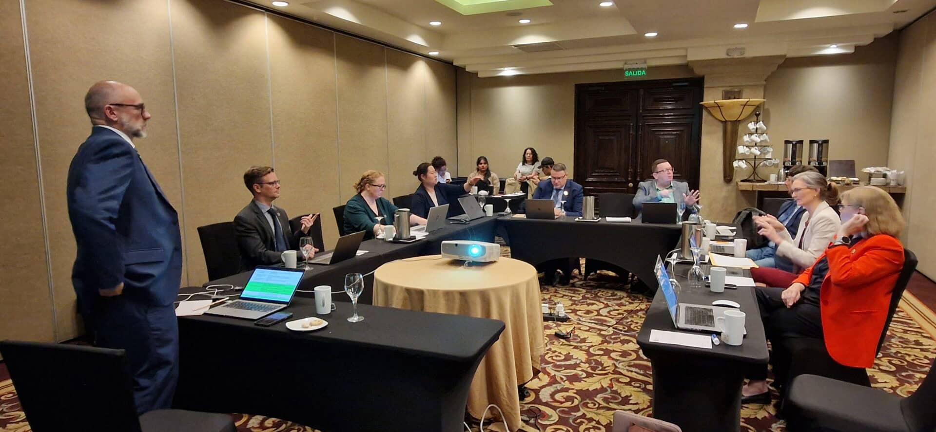 A group of people in business attire are seated around a U-shaped conference table with laptops and papers, having a meeting in a hotel conference room.