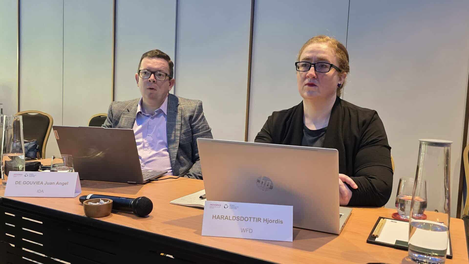 Two people sit at a conference table with laptops open in front of them, participating in a meeting. Nameplates and water glasses are visible on the table.