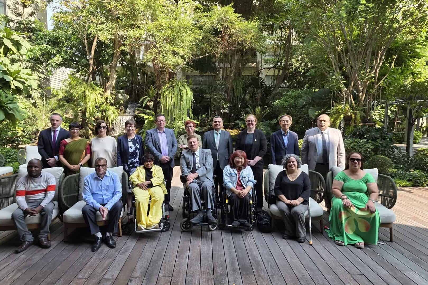 A group of people, some seated and some standing, pose for a photo outdoors on a wooden deck surrounded by greenery.
