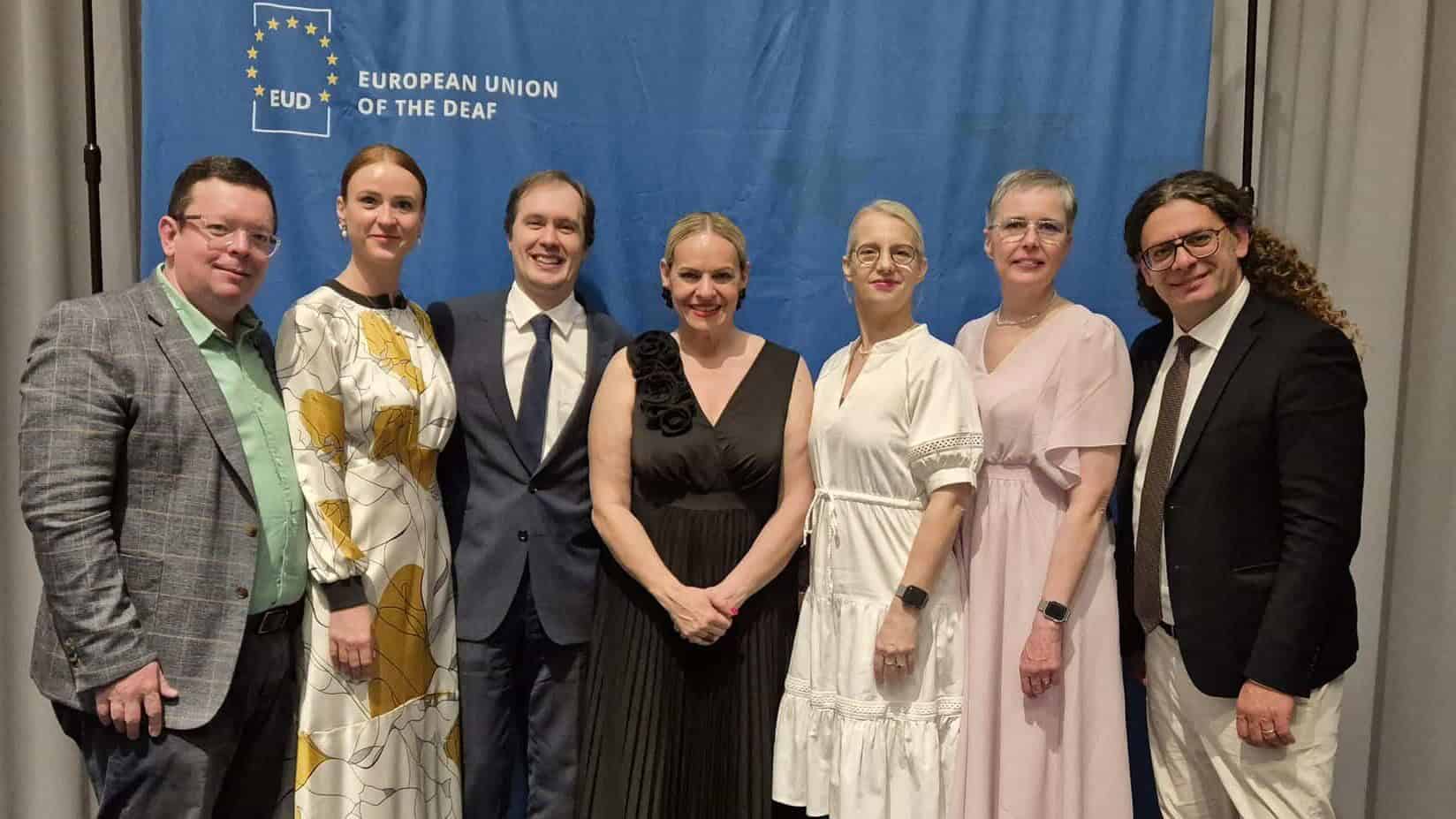 Seven people stand side by side in formal attire in front of a blue European Union of the Deaf backdrop.