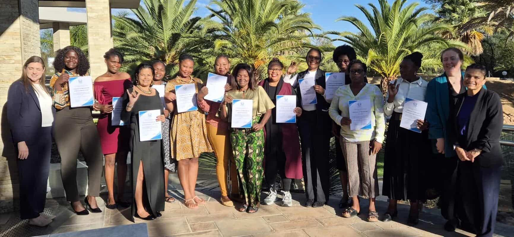 A group of people stand outdoors holding certificates, posing for a group photo on a sunny day with palm trees and blue sky in the background.
