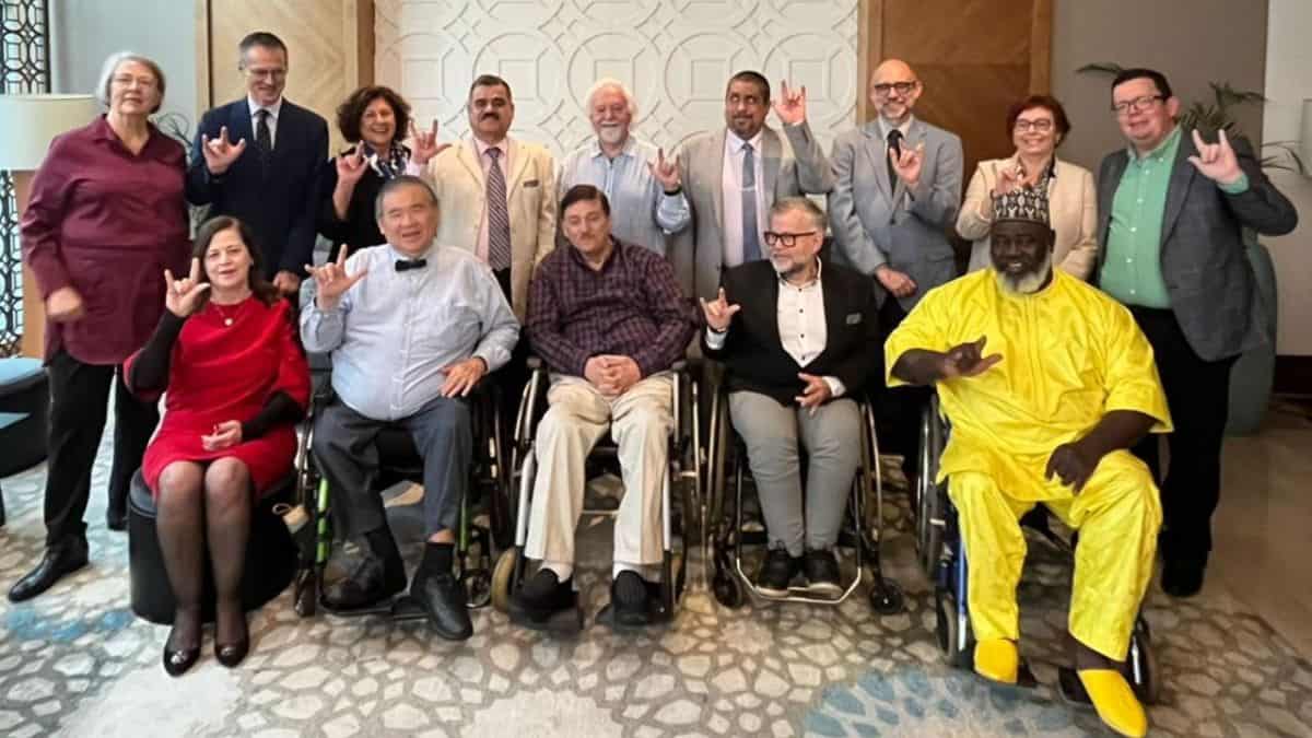 A group of people, including several in wheelchairs, pose for a photo indoors. Some are making a hand gesture, and one is wearing a yellow outfit. The background features a patterned wall.