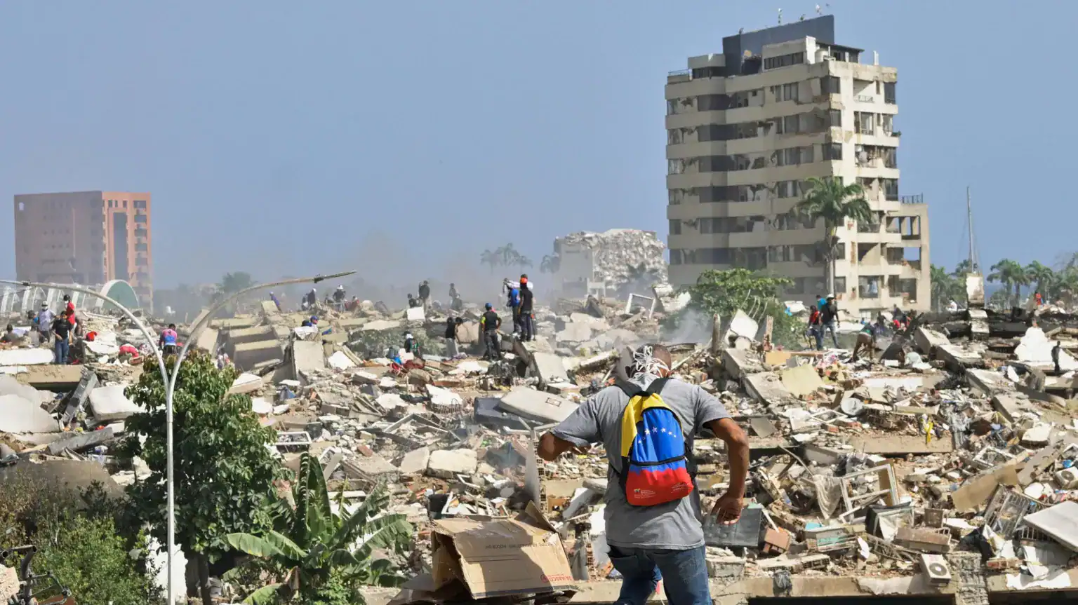 People walk among debris and collapsed buildings in an urban area, with one partially damaged building standing in the background under a clear sky.