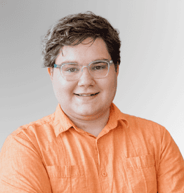 Person with short brown hair and glasses wearing an orange shirt, smiling and standing in front of a light grey gradient background.