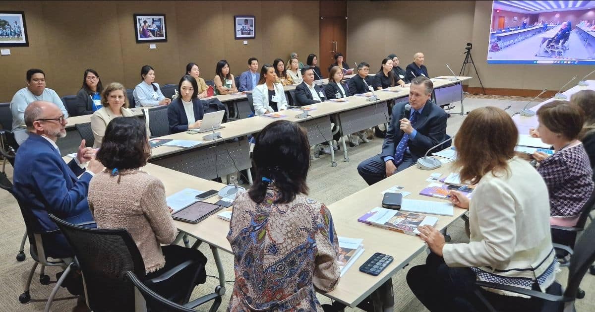 A group of people in a conference room engage in a meeting, seated around tables with laptops and documents. A presenter speaks into a microphone facing the attendees.