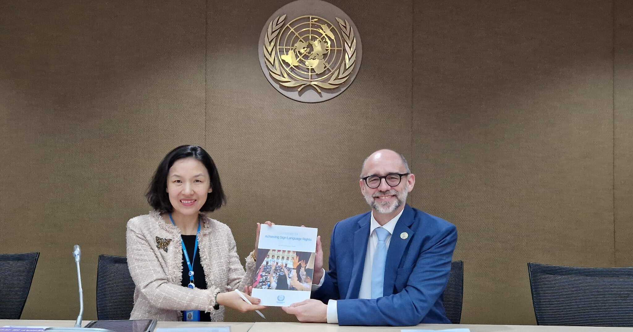 Two people smiling and holding a document together at a conference table, with a United Nations emblem on the wall behind them.