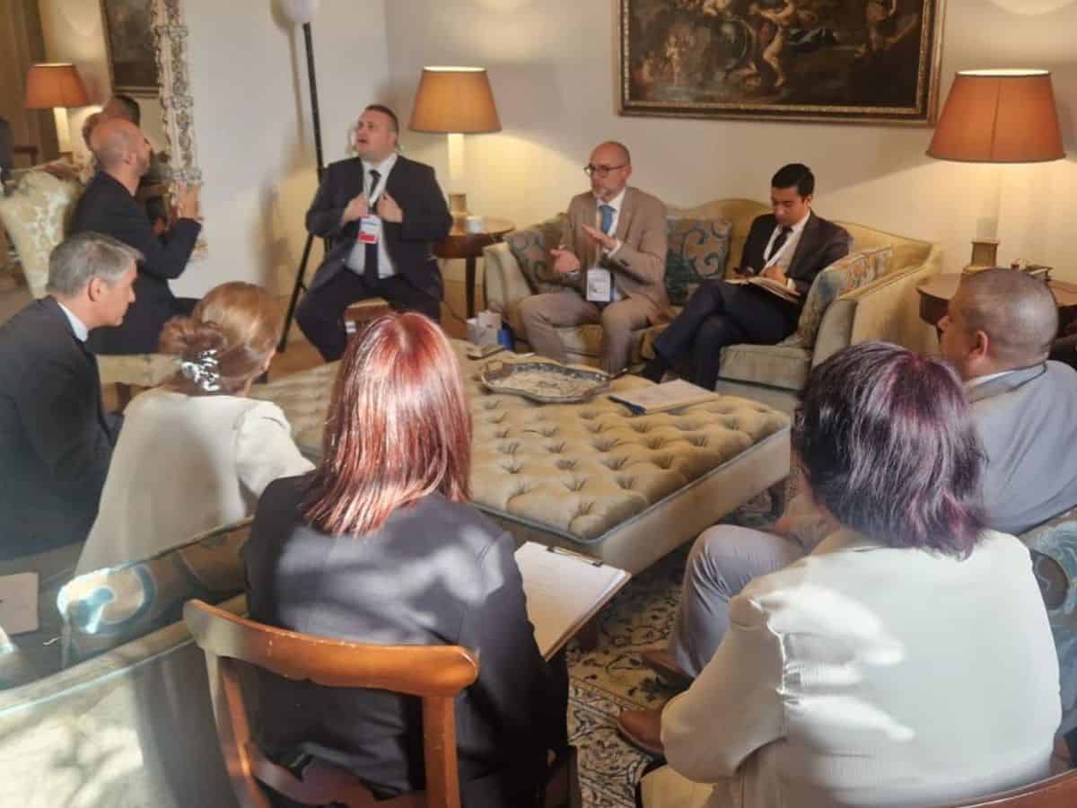 A group of people in formal attire sit in a living room during a meeting. Some are taking notes, and two lamps light the room.