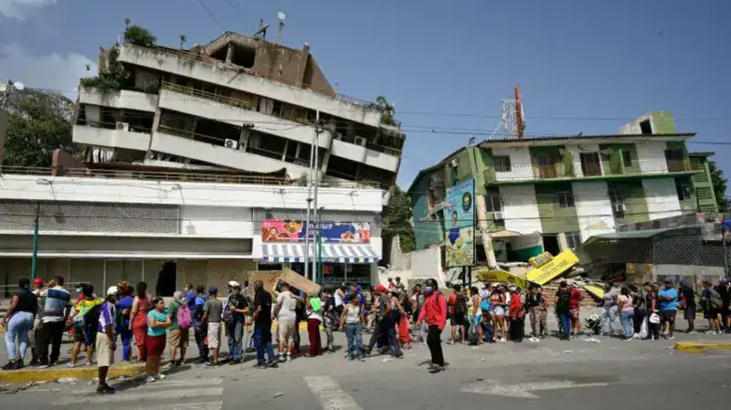 People stand in a long queue near damaged buildings, one of which is tilted and leaning, likely due to an earthquake.
