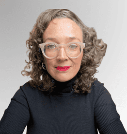 A woman with curly brown hair, wearing clear glasses, red lipstick, and a black polo neck, sits in front of a plain light grey background.