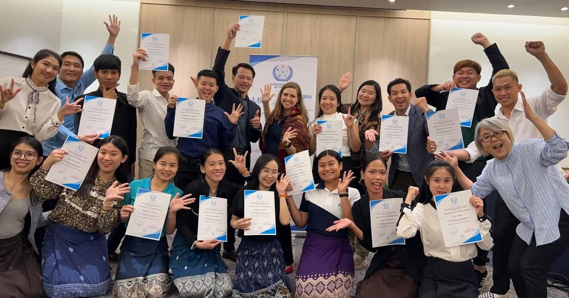 A group of people pose indoors, holding certificates and smiling enthusiastically, with a banner in the background.