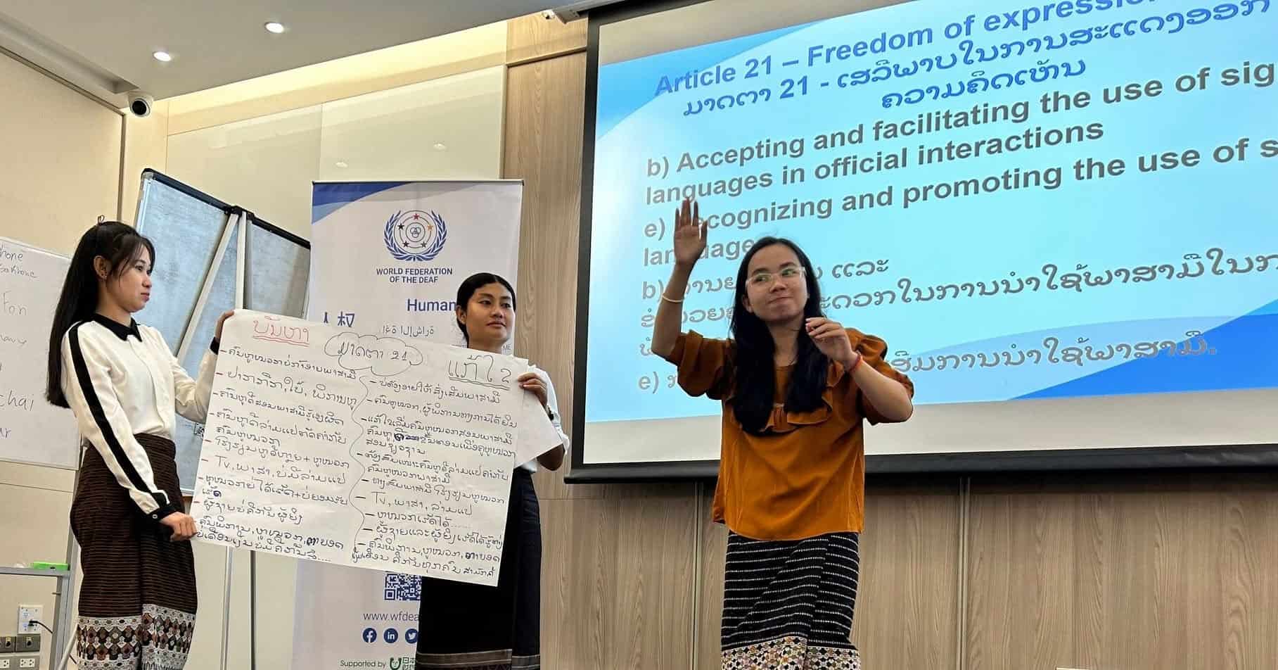 Three women present information in front of a projected screen and a poster board, discussing Article 21 on freedom of expression at an indoor event.