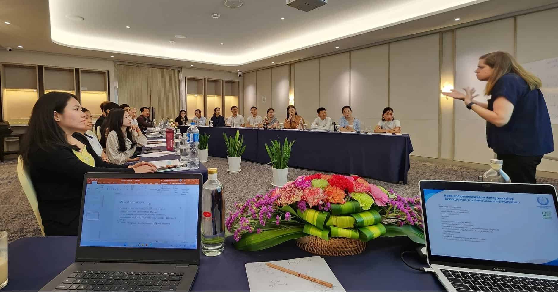 A woman stands and gestures whilst presenting to a group seated around U-shaped tables in a conference room; laptops and documents are visible in the foreground.