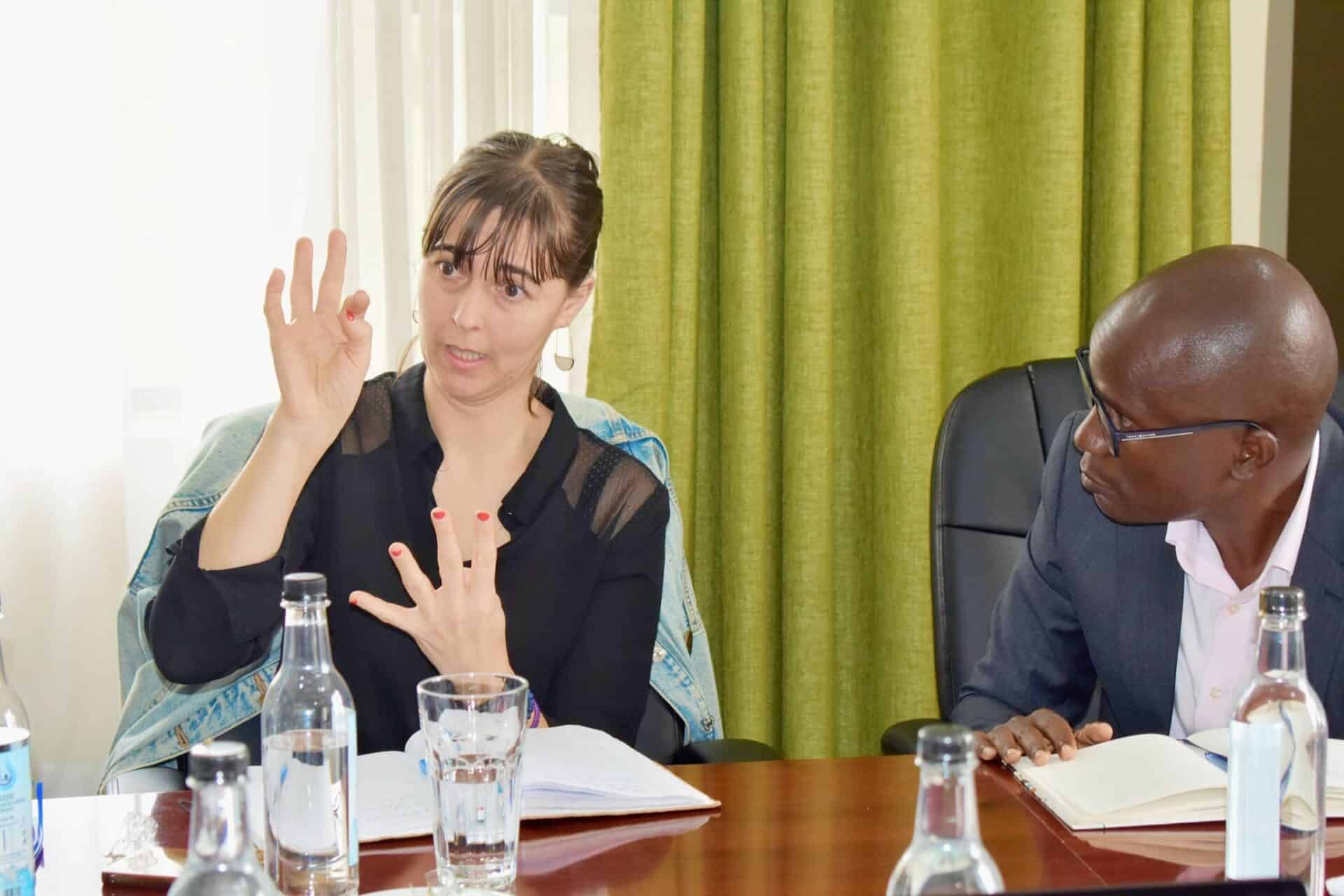 A woman gestures with her hand while speaking at a meeting table with notebooks, glass bottles, and a man listening beside her.