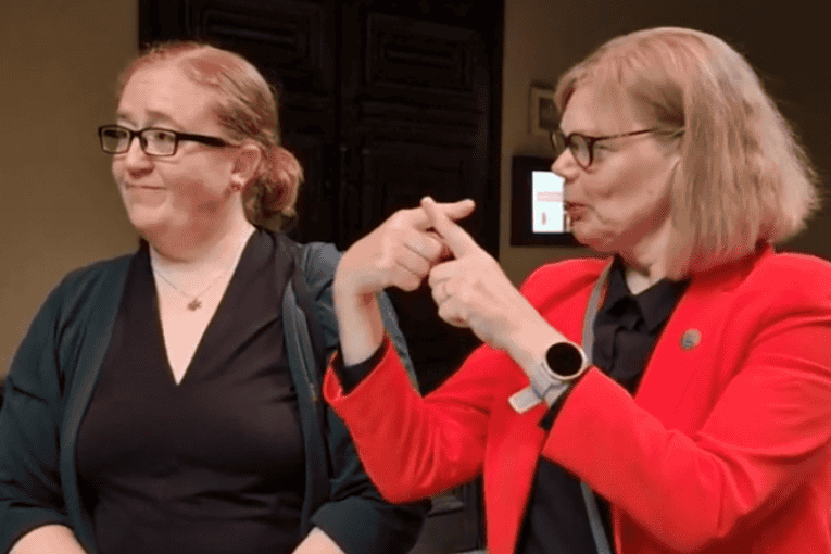 Two women stand at a round table; one writes in a notebook while the other gestures with her hands as if signing.