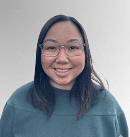 A person with straight, dark hair and glasses is smiling at the camera, wearing a dark green jumper against a light grey background.