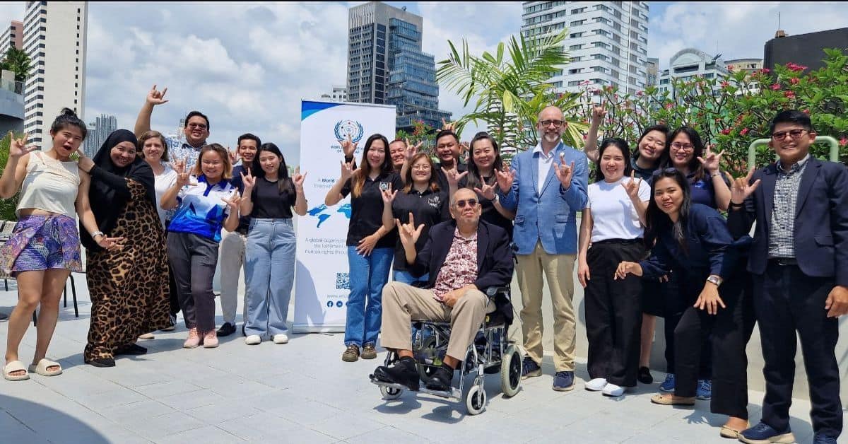 A diverse group of people, including a person in a wheelchair, pose outdoors in front of a banner, with buildings and trees in the background.