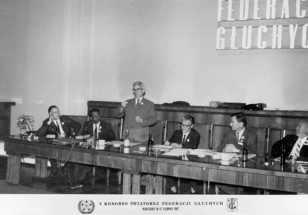 A man speaks at a table with five seated individuals at a conference. A banner reads "Federacja Głuchych.