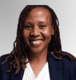 A woman with long, plaited hair smiles at the camera, wearing a dark blazer over a white top, against a gradient grey background.