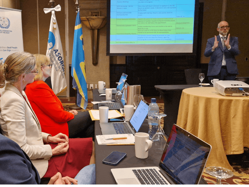 Three people sit at a table with laptops during a meeting while a man stands and presents slides on a screen at the front of the room.