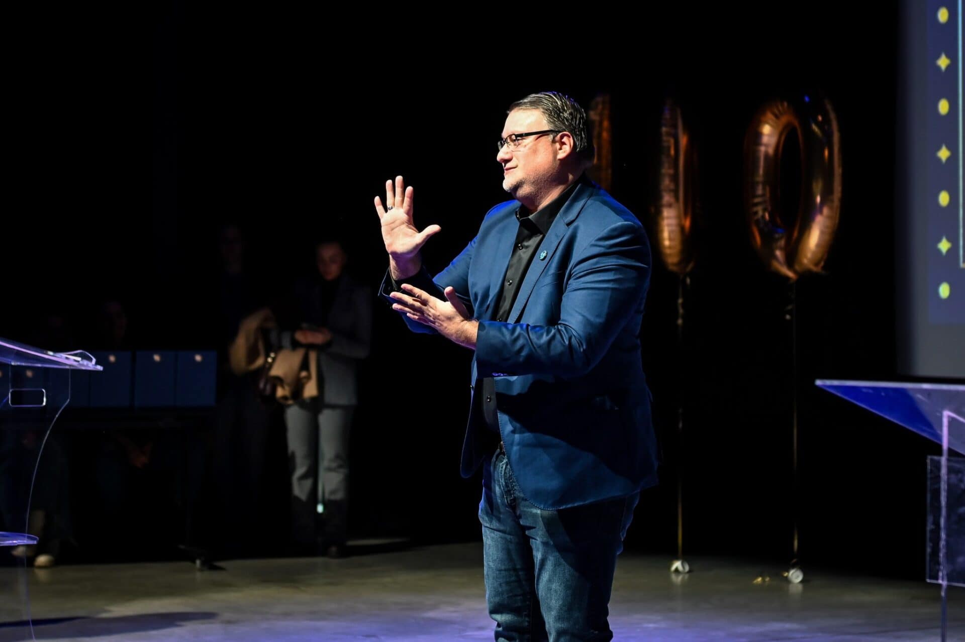 A man in a blue blazer gestures whilst speaking on stage, with gold balloons and a projection screen visible in the background.
