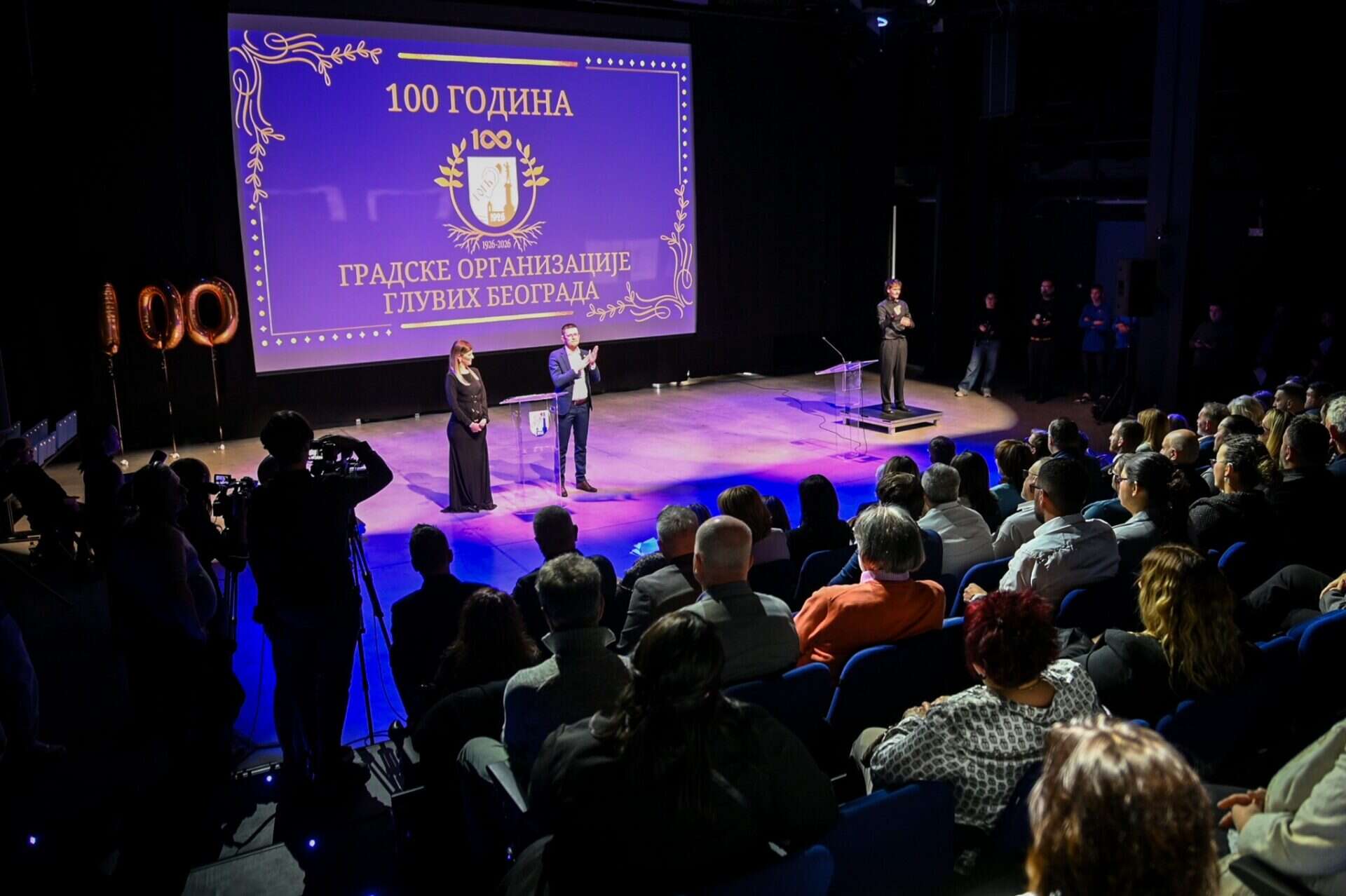 A man speaks on stage at an event celebrating the 100th anniversary of the Belgrade Association of the Deaf, with an audience seated and a sign language interpreter present.