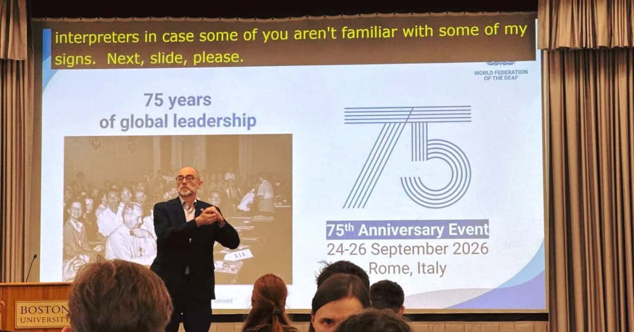 A man uses sign language on stage at a Boston University event, with a presentation slide behind him about the World Federation of the Deaf’s 75th Anniversary Event in 2026.