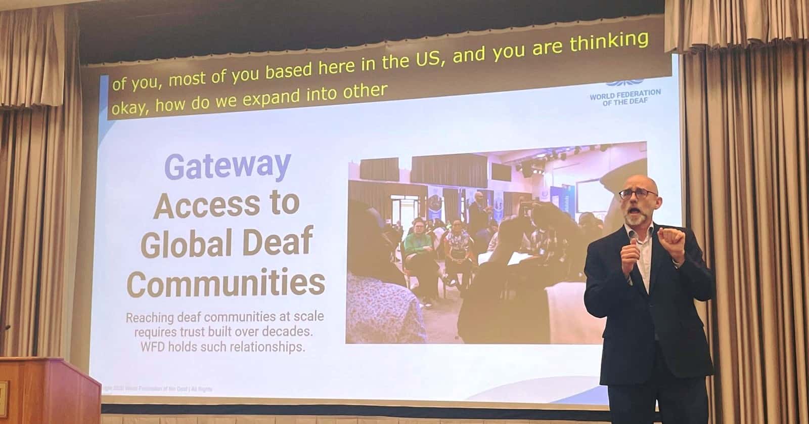 A man in a suit signs during a presentation on "Access to Global Deaf Communities" at a conference. A projected slide and subtitles are visible behind him.