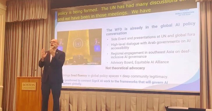 A man in a suit gestures whilst speaking onstage at a Boston University event; a presentation slide about AI policy is displayed behind him.