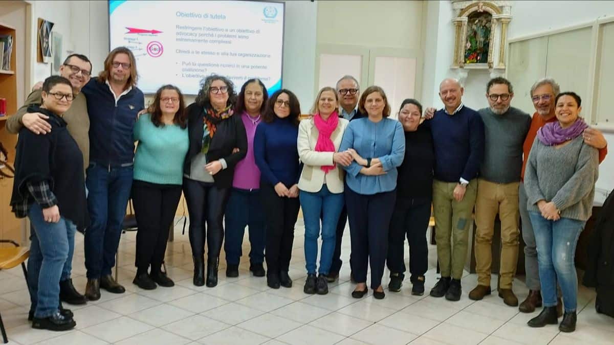 A group of people in a classroom setting, standing close together and smiling at the camera. A presentation slide is visible in the background.