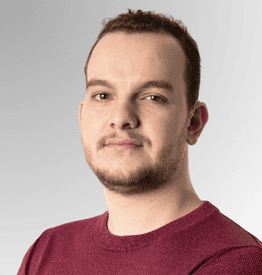 A man with short brown hair and a trimmed beard wearing a maroon jumper stands against a light grey background, looking at the camera with a neutral expression.