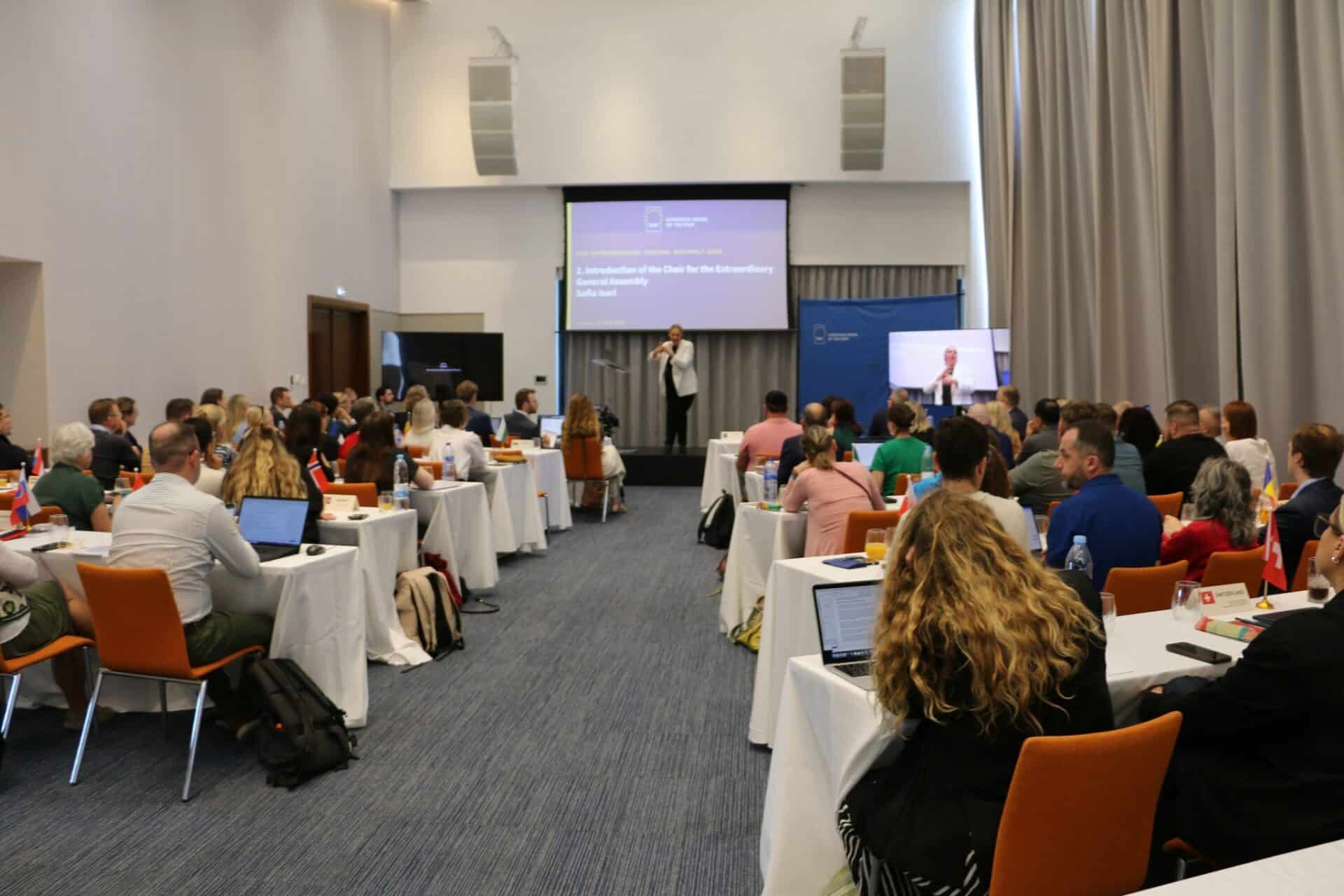 A group of people seated at tables in a conference room listens to a presenter speaking at the front with a projected screen behind them.