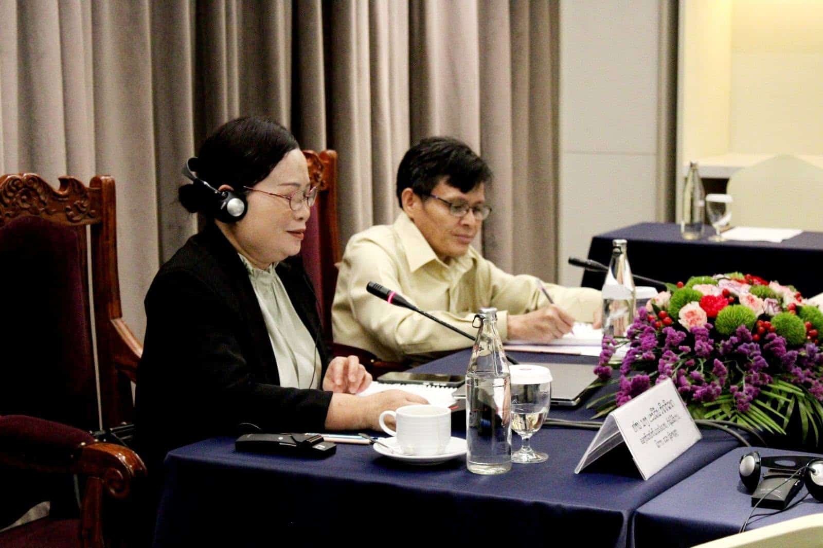 Two people sit at a conference table with microphones, papers, and flowers. One person is speaking and wearing a headset; the other takes notes.