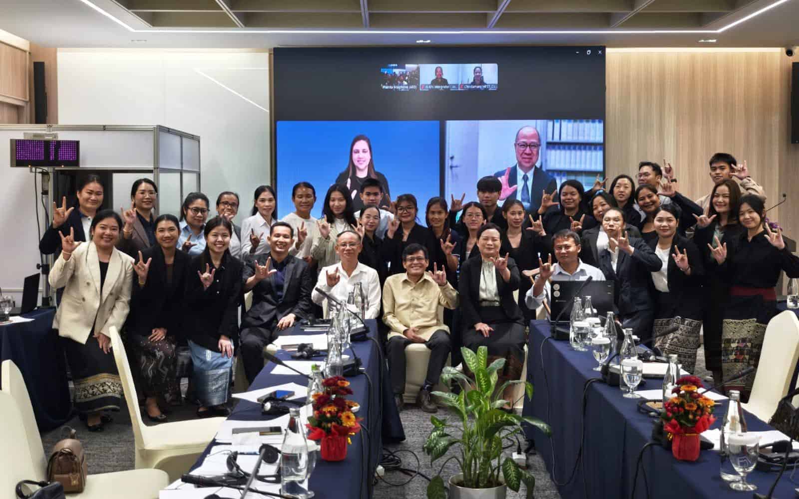 A large group of people pose for a photo in a conference room, with three individuals visible on a screen behind them during a video call. Many people are making hand signs.