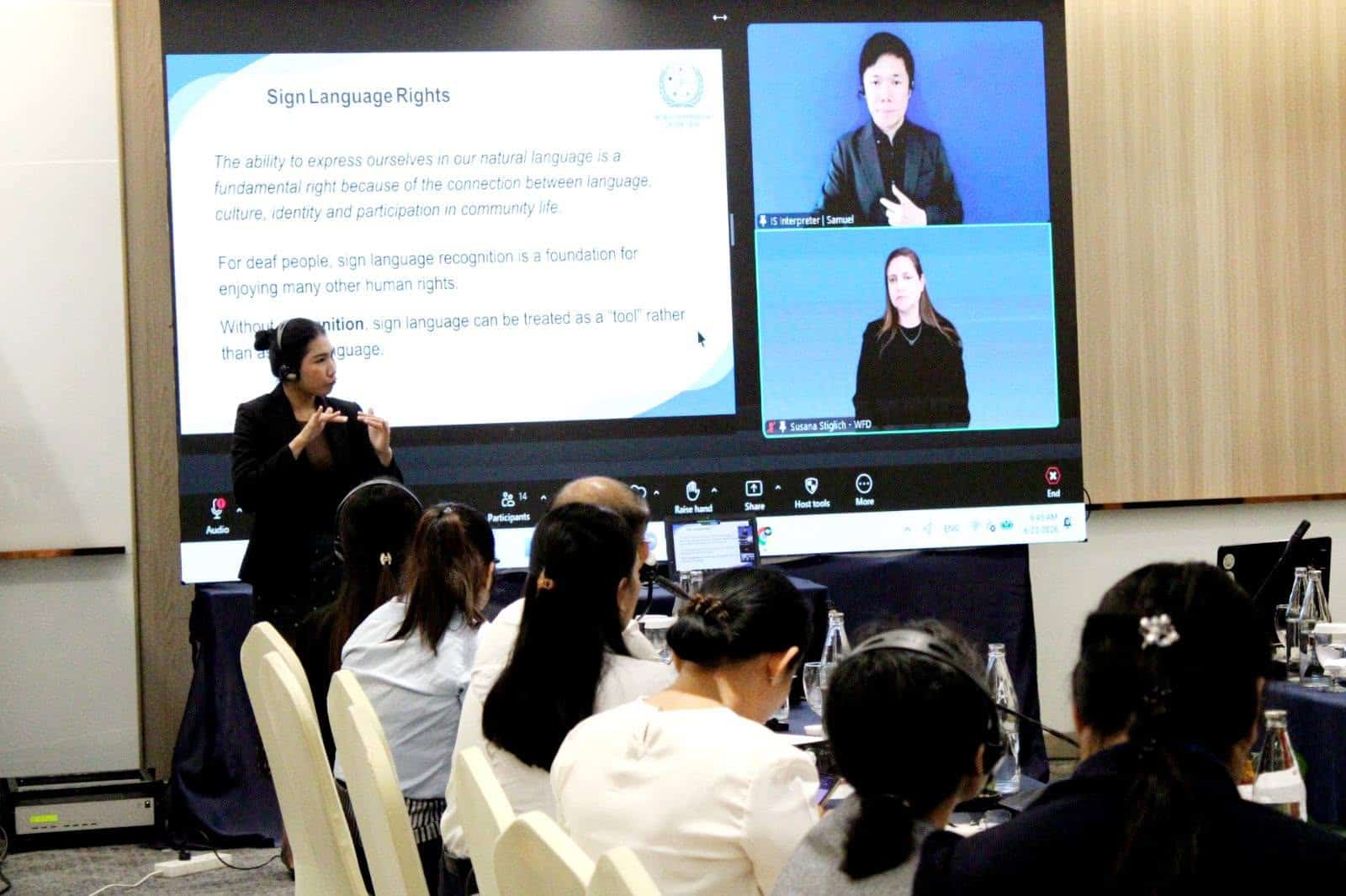 A presenter uses sign language in front of an audience, whilst a screen displays a slide on sign language rights and two video sign language interpreters.