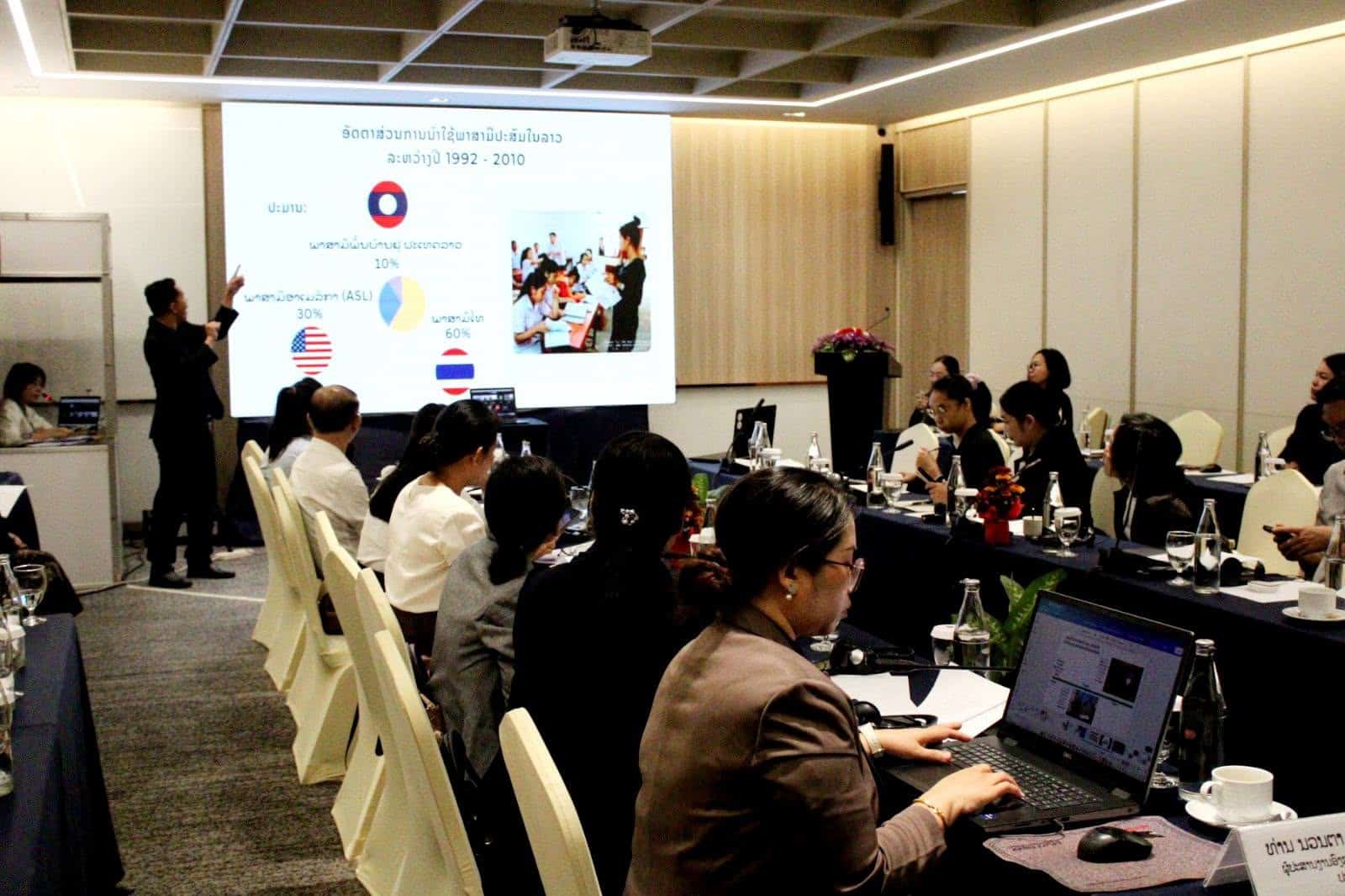 A group of people in a conference room listen to a presenter showing a projected slide with charts and images.