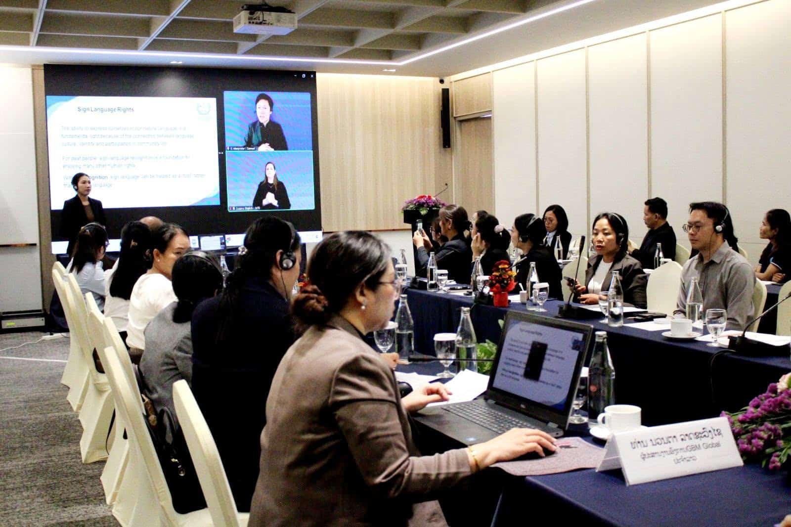 A group of people attend a conference in a meeting room, with a presenter speaking and sign language interpreter displayed on a screen at the front.