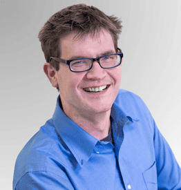 Smiling man with short brown hair and glasses wearing a blue button-up shirt against a grey gradient background.